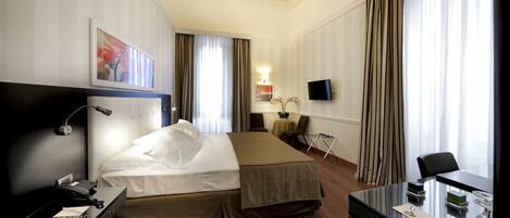 A hotel room with a bed, a desk with a telephone, a television, and a small table with a vase of flowers.