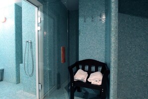 A bathroom with a glass shower enclosure, a wooden bench with towels, and a tiled wall. - Il Principe Hotel (Catania)