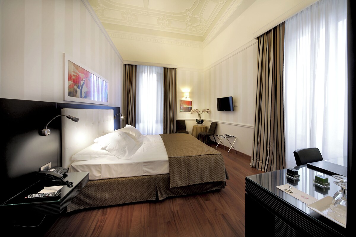 A hotel room with a bed, bedside table, lamp, TV, and a desk with a chair.