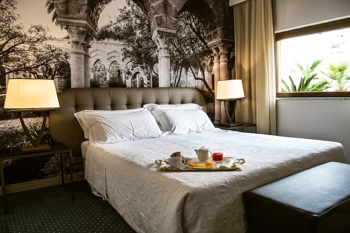 A hotel room with a large bed, two bedside lamps, a tray with breakfast, and a mural of trees and architecture on the wall.