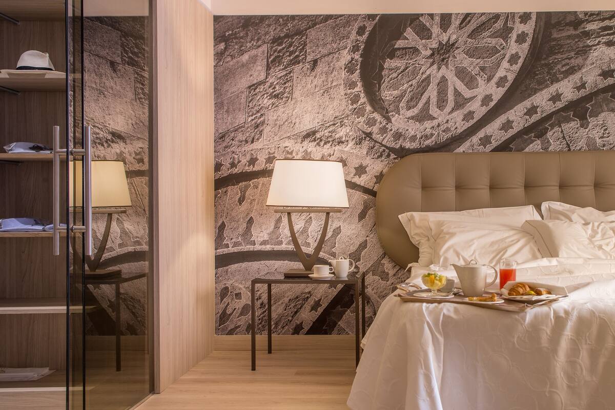 A hotel room with a bed, bedside table with a lamp, a tray with breakfast items, and a decorative wall mural.