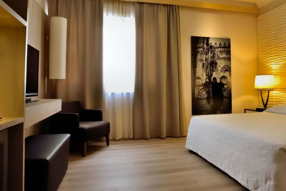 A hotel room with a bed, a desk, a chair, a large window with curtains, a wall-mounted lamp, and a wall art piece.