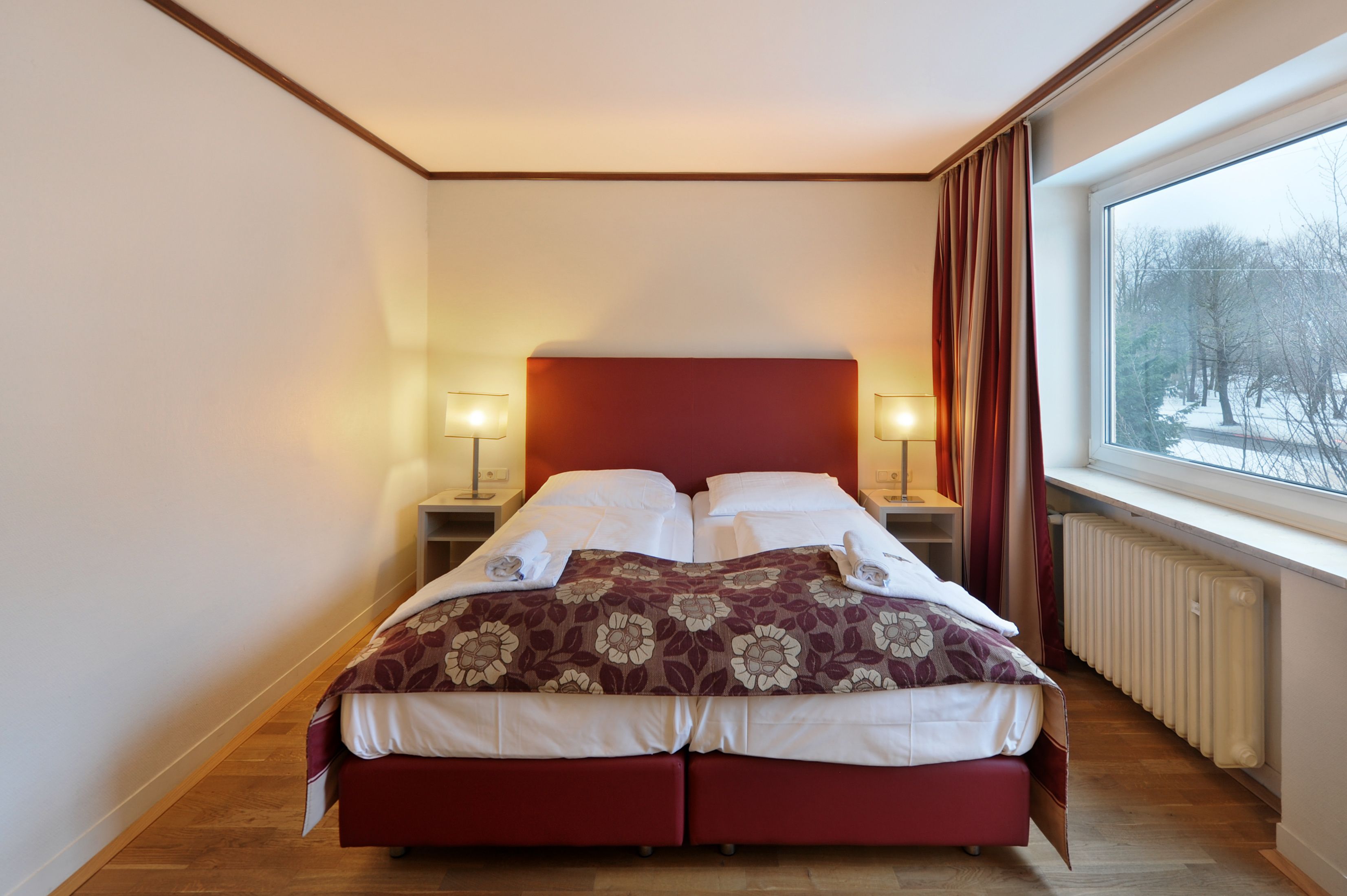 A double bed with a red headboard, two bedside tables with lamps, a window with a view of trees, and a radiator.