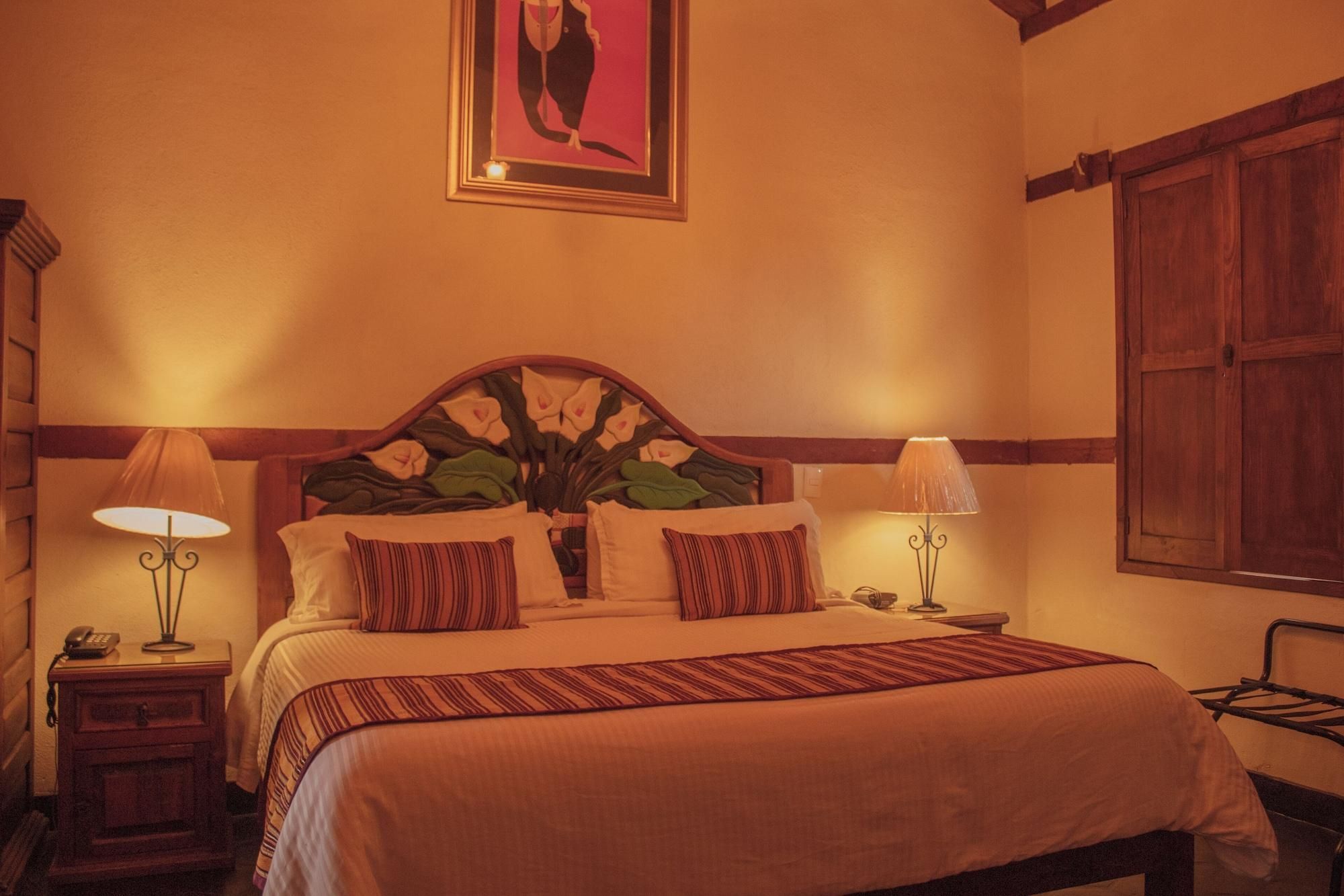 A hotel room with a bed, two bedside lamps, a nightstand with a phone, a framed picture on the wall, and a wooden door.