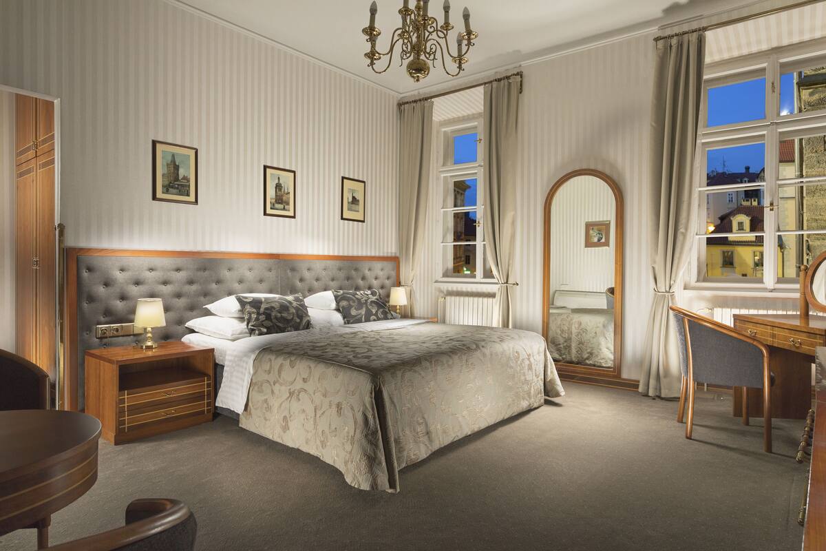 Quarto com cabeceira estofada, mesa de cabeceira de madeira, escrivaninha e cadeira. Há três quadros na parede, uma janela com cortinas e vista para edifícios.