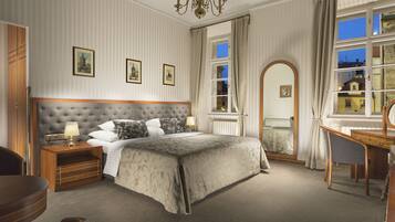 Un dormitorio con cabecera capitonada, una mesita de noche de madera, un escritorio y una silla. Hay tres cuadros enmarcados en la pared, una ventana con cortinas y vistas a edificios.