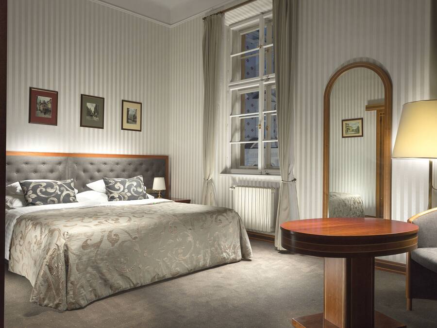A hotel room with a large bed, a round wooden table, a chair, a window with curtains, and framed pictures on the wall.