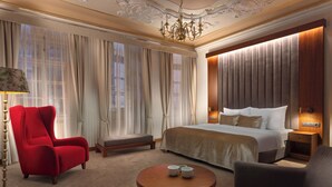Una camera d'albergo con un letto grande, una poltrona rossa, un tavolino rotondo in legno e un lampadario.