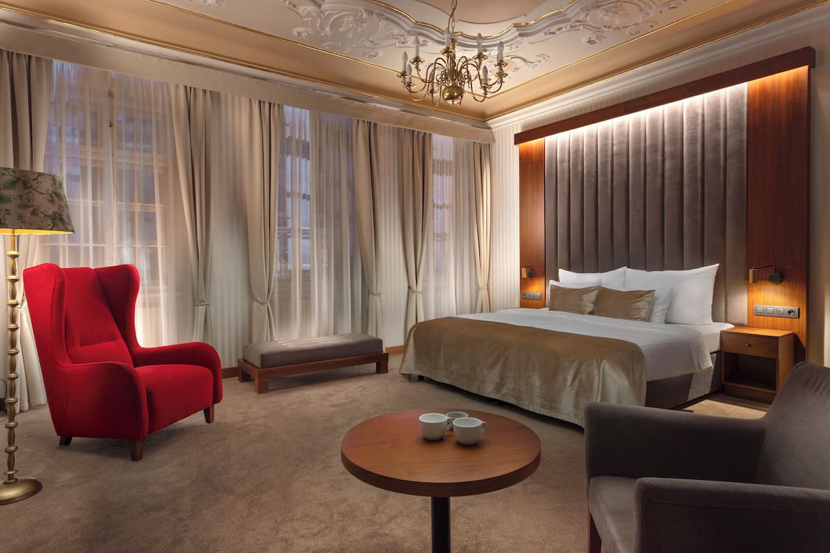 Quarto de hotel com uma cama grande, uma poltrona vermelha, uma mesa de madeira redonda e um candelabro.