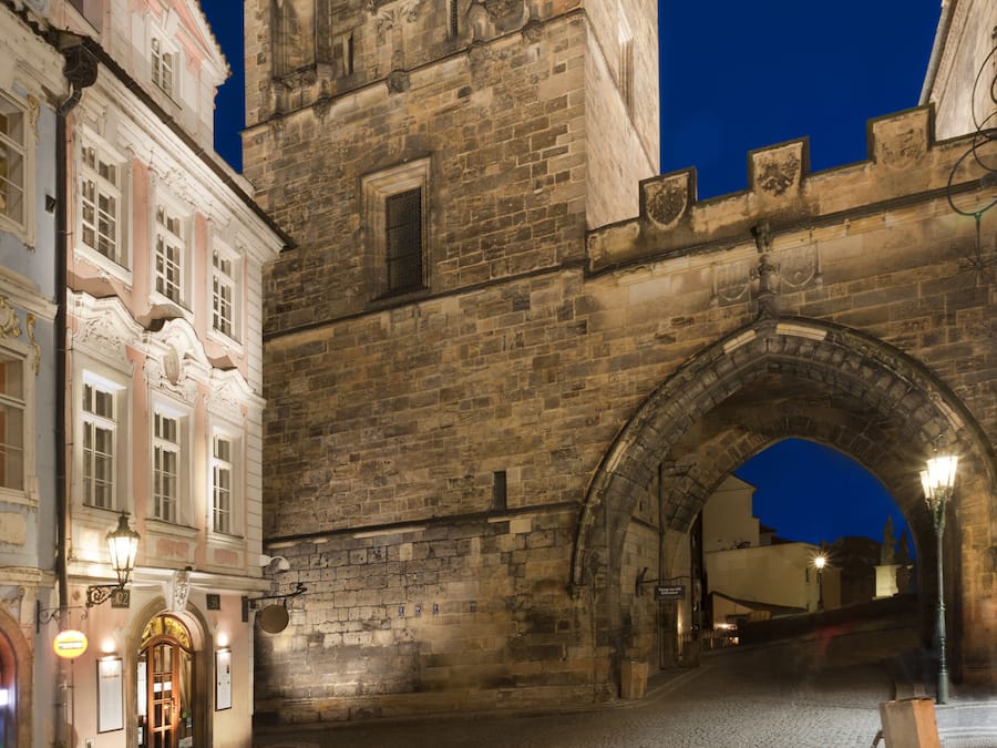 Un puente de piedra histórico con un arco, calle adoquinada y edificios tradicionales con letreros iluminados.
