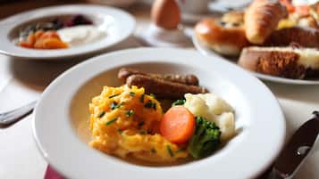 Una mesa de comedor con platos de comida que incluyen huevos revueltos, salchichas, puré de patatas y verduras.