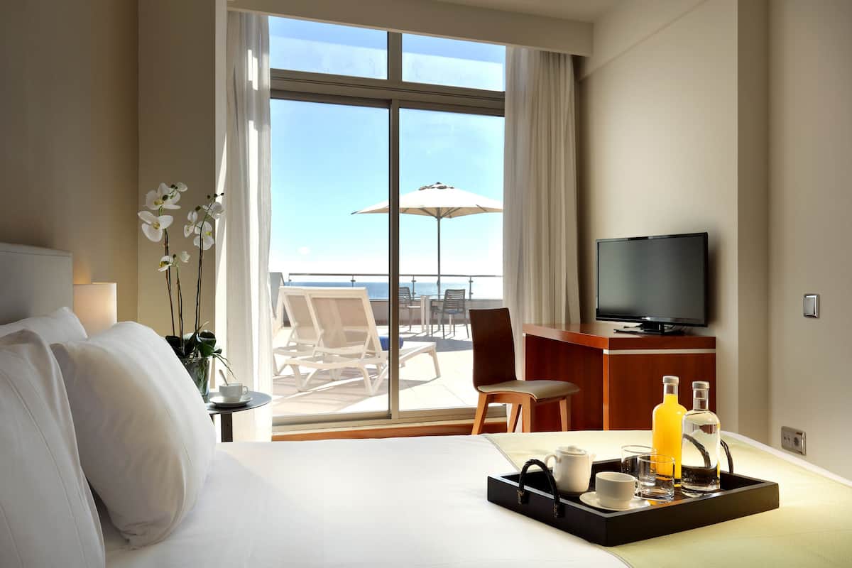 Ein Hotelzimmer mit Bett, Fernseher, Tablett mit Getränken und Tassen, Stuhl und Blick auf das Meer durch große Fenster.