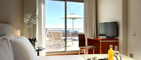 A hotel room with a bed, a TV, a tray with drinks and cups, a chair, and a view of the ocean through large windows.