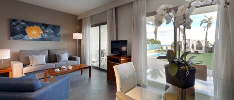 A modern hotel room with a sofa, a coffee table, a TV, and a glass table with chairs. There is a large window with curtains and a view of the outdoors.