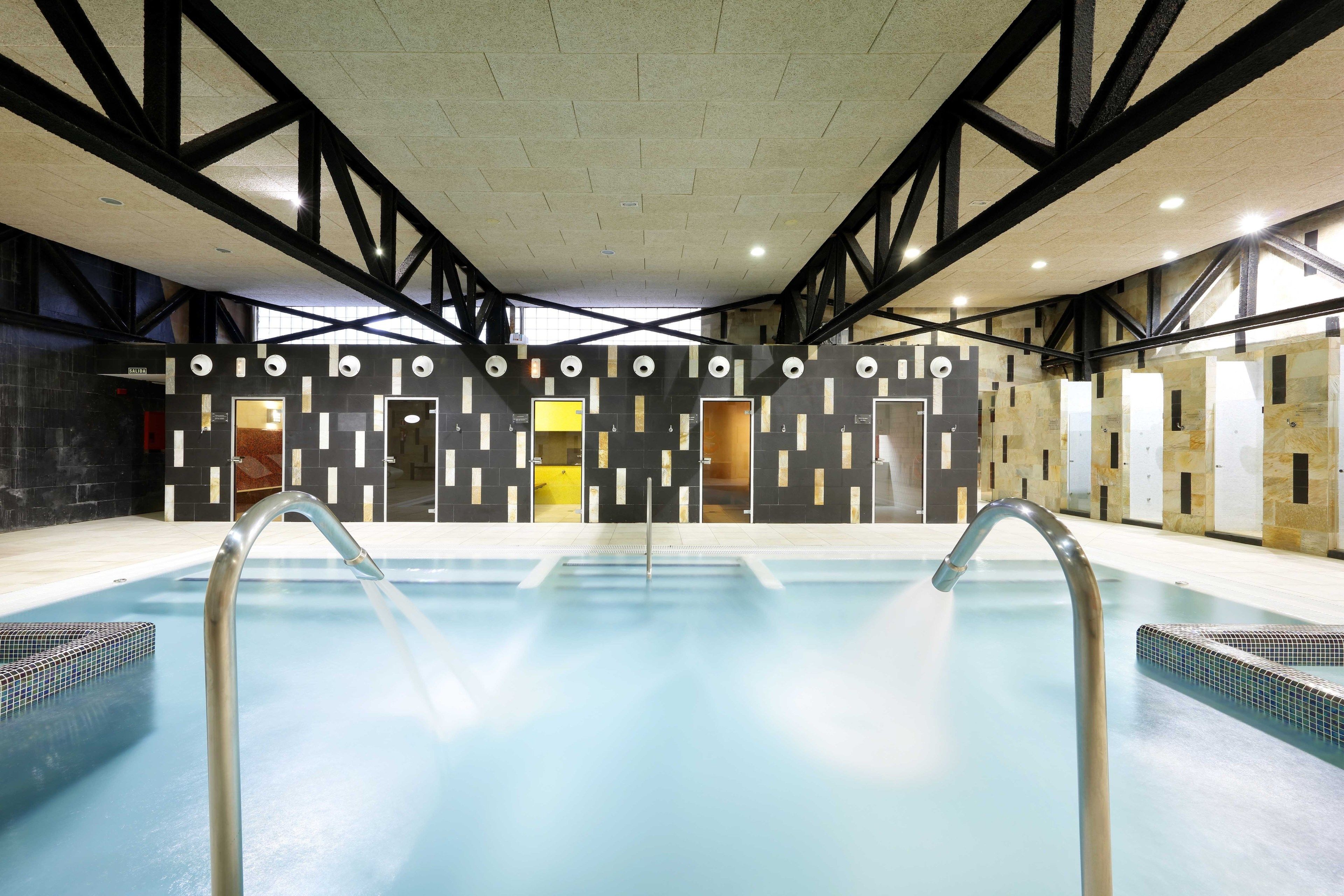Una piscina con diseño arquitectónico moderno, que destaca por su techo alto y vigas estructurales negras.