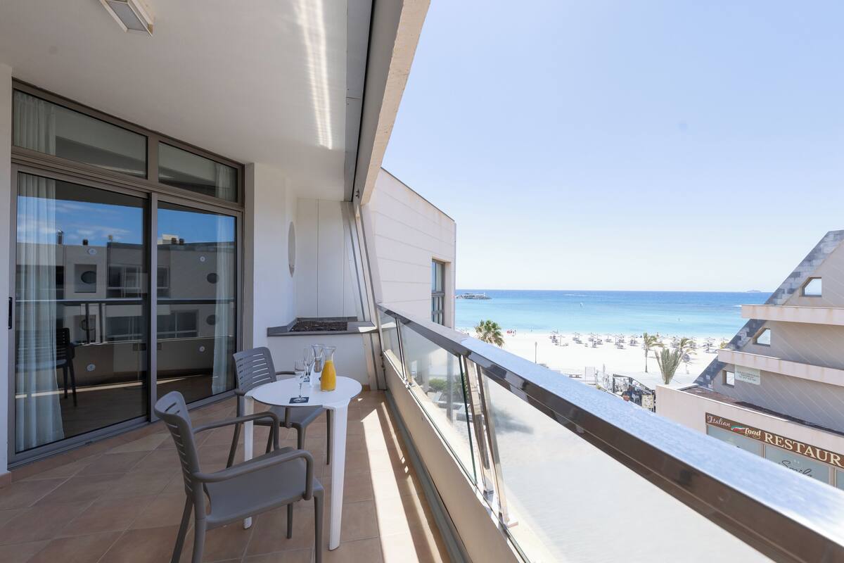 Ein Balkon mit Tisch und Stühlen, der Blick auf einen Strand und ein Restaurant bietet.