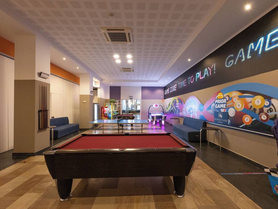 A pool table in a hotel lobby with a neon sign reading "GAME TIME TO PLAY!" and a colorful mural with pool balls.