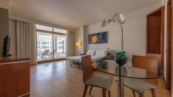 Quarto de hotel com cama, escrivaninha, mesa de vidro e uma cadeira. Há uma janela com cortinas, um quadro na parede e uma planta sobre a mesa.
