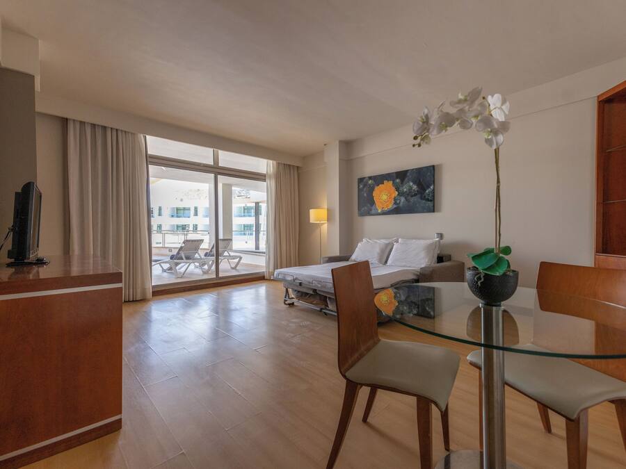 Habitación de hotel con cama, escritorio, mesa de vidrio y una silla. Hay una ventana con cortinas, un cuadro en la pared y una planta sobre la mesa.
