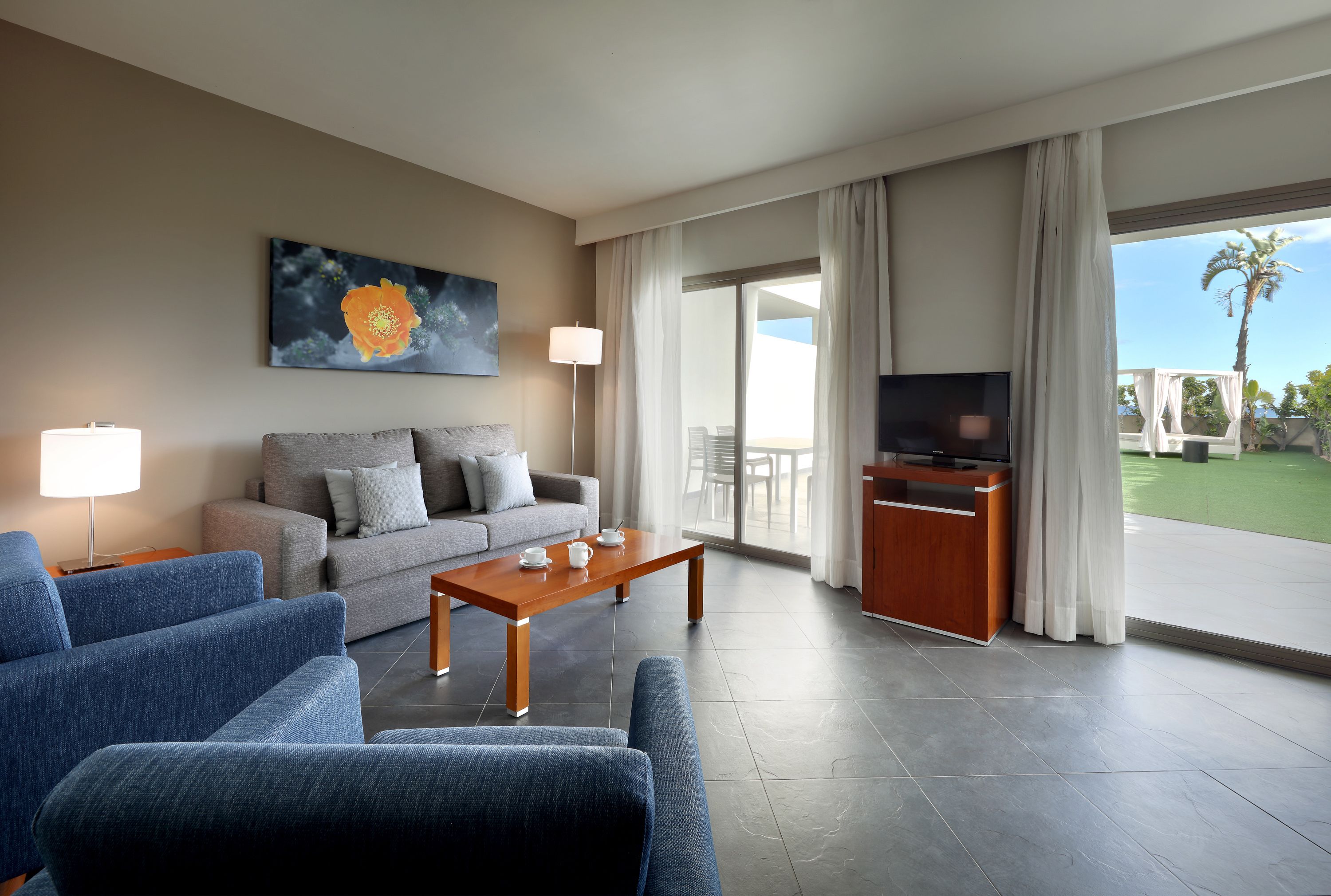 Una sala de estar moderna con un sofá gris, sillones azules, una mesa de centro de madera, un televisor de pantalla plana y un ventanal con cortinas.
