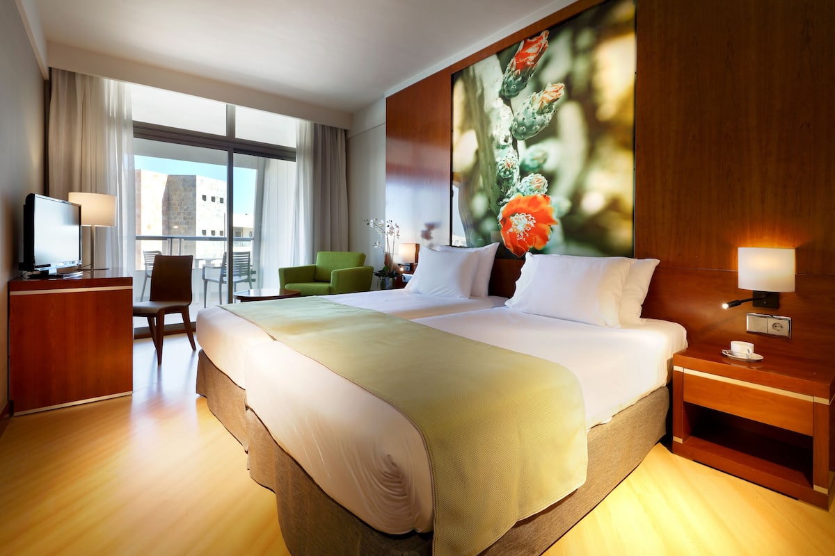 Ein Hotelzimmer mit einem großen Bett, einem Schreibtisch mit Computer, einem grünen Sessel und einem großen Fenster mit Blick auf die Stadt.