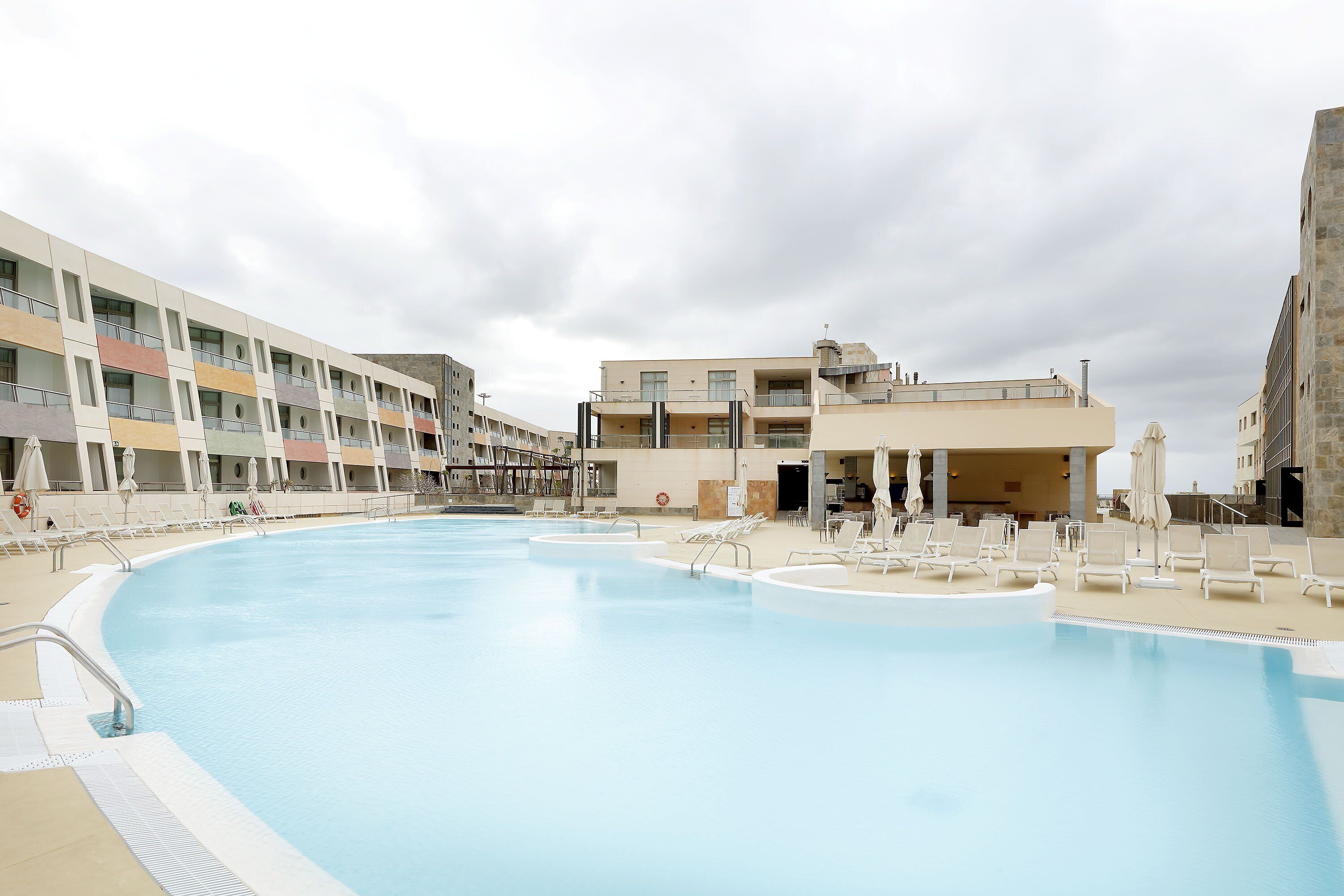 A swimming pool with lounge chairs and umbrellas, surrounded by multi-story buildings with balconies.