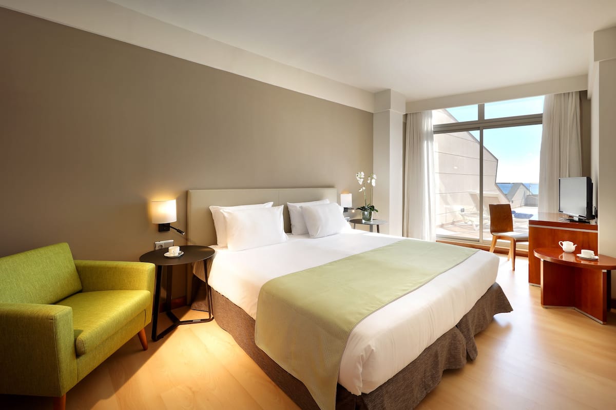 Ein Hotelzimmer mit einem großen Bett, einem grünen Sessel, einem Schreibtisch mit Stuhl und einem großen Fenster mit Blick.