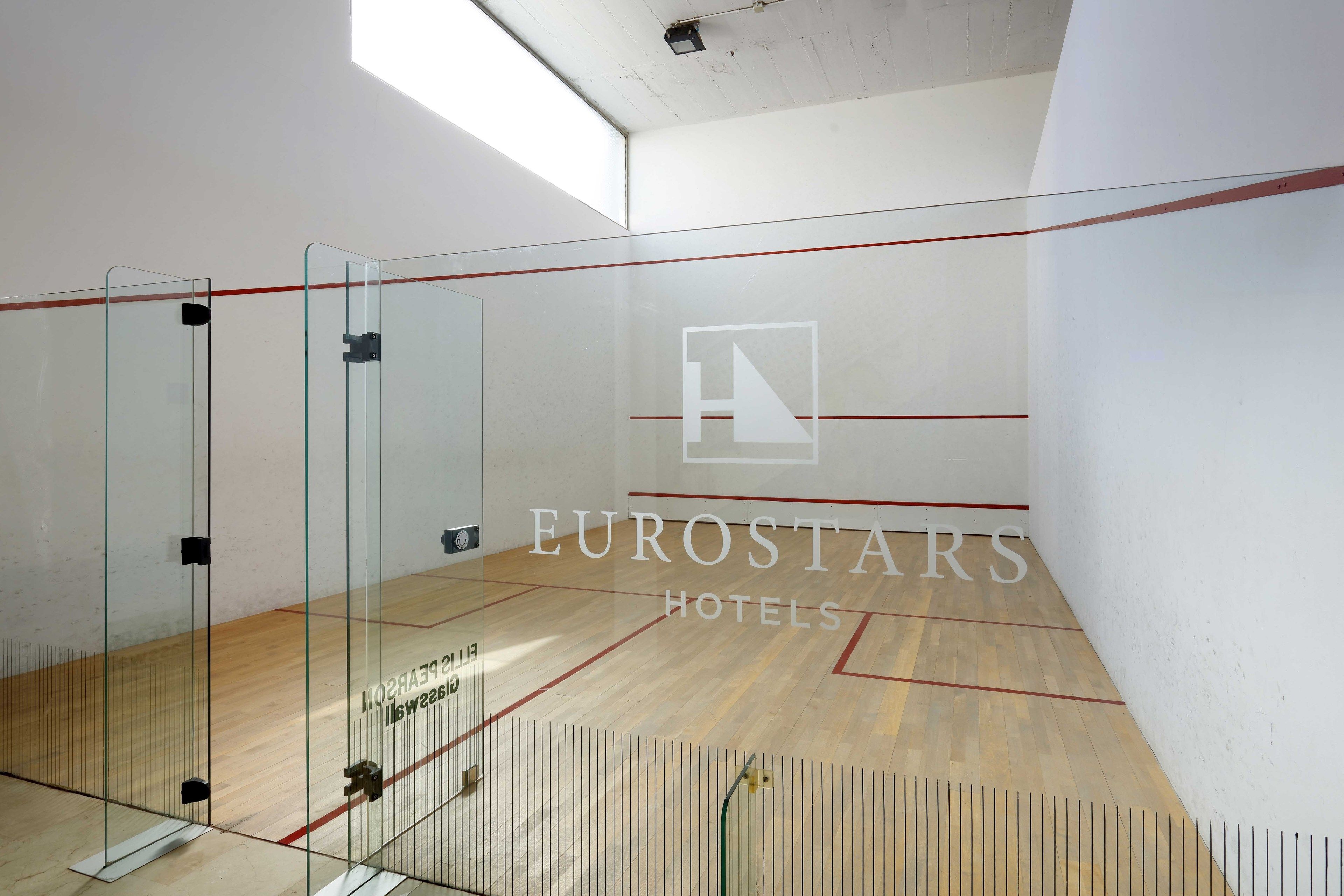 Un court de squash avec des murs en verre, un plancher en bois et le logo de la chaîne hôtelière Eurostars.