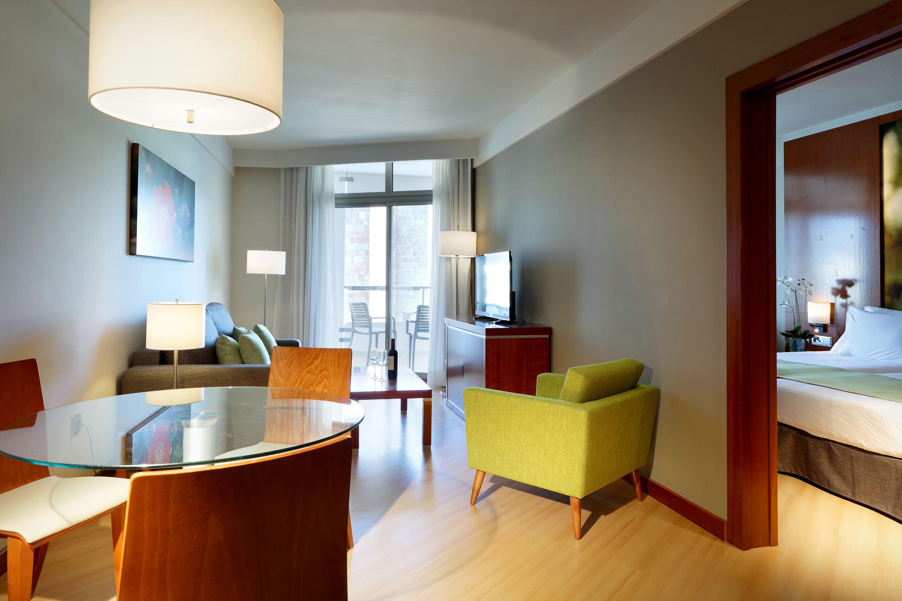 Habitación de hotel con mesa de vidrio, sofá, cama, televisión y vista al exterior.