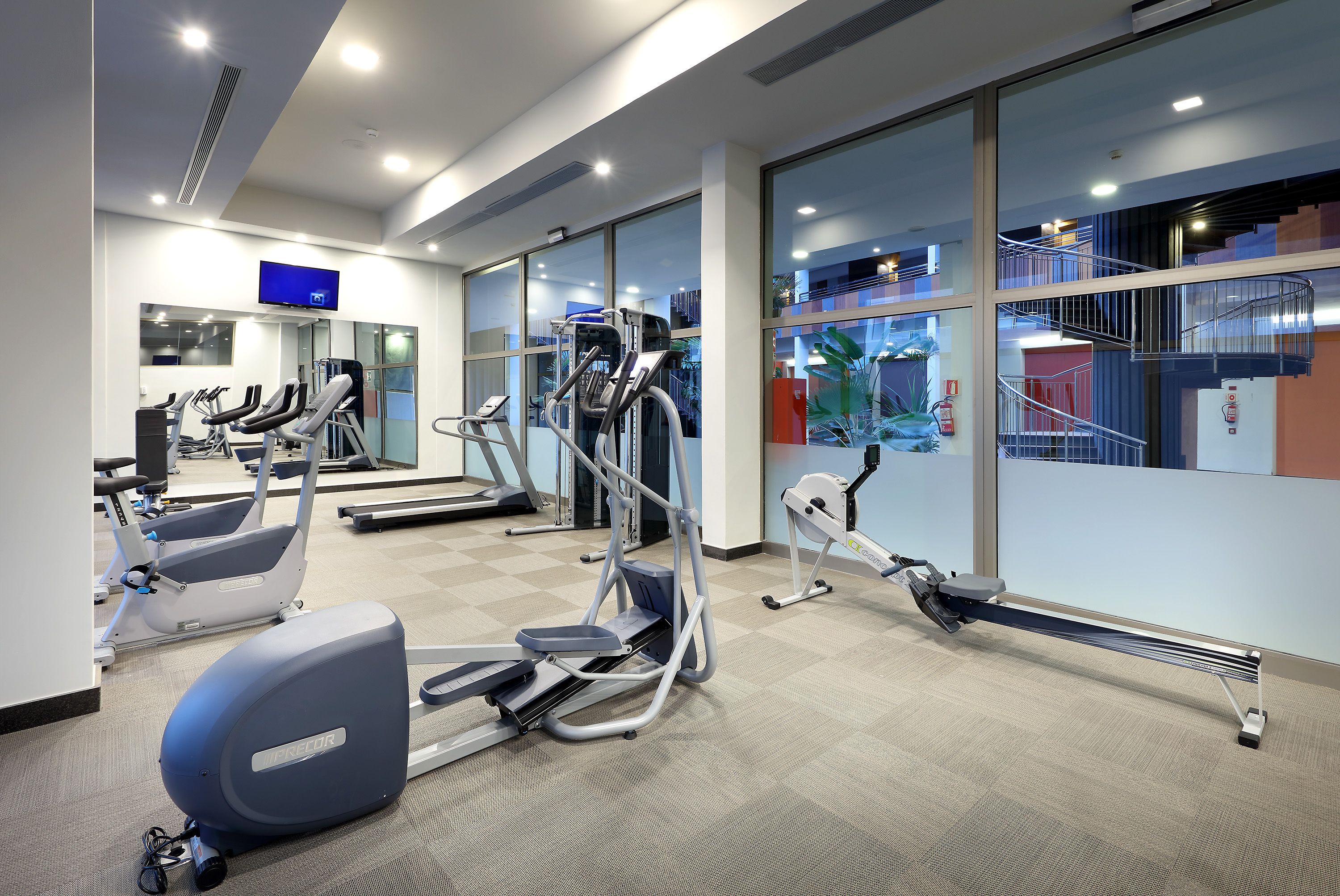 Ein modernes Fitnessstudio mit unterschiedlichen Trainingsgeräten, großen Fenstern und einer sichtbaren Treppe.