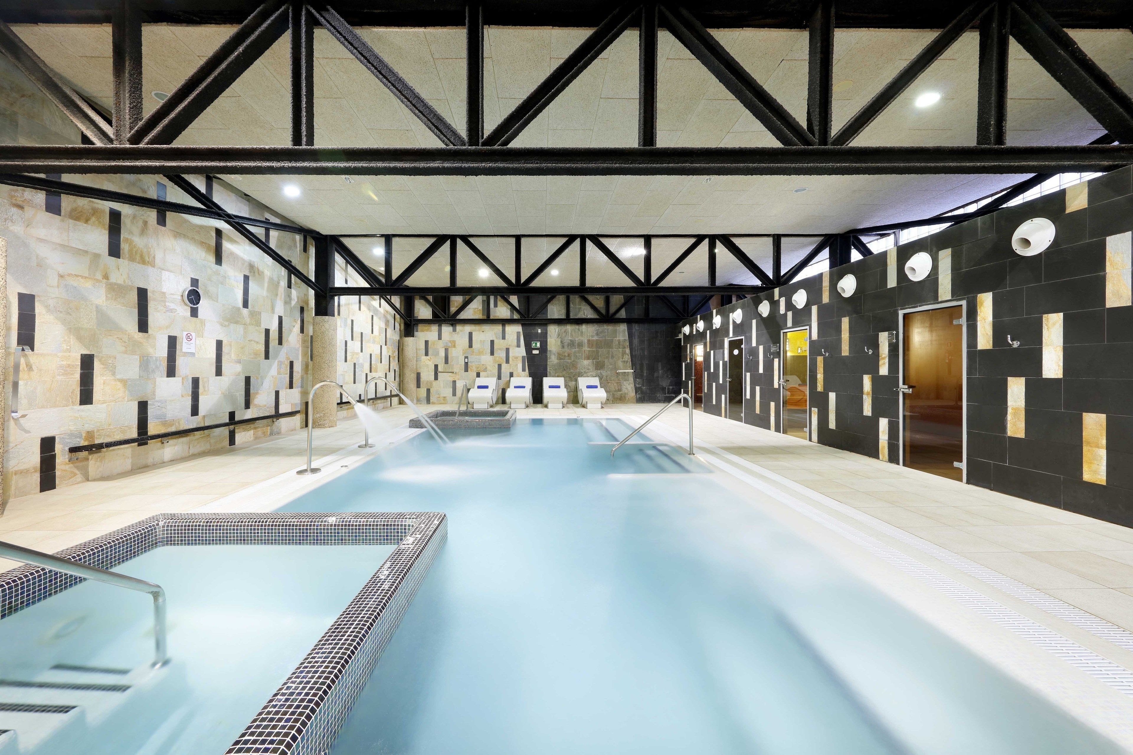 An indoor swimming pool with a modern design, featuring a geometric wall pattern and a high ceiling with truss beams.