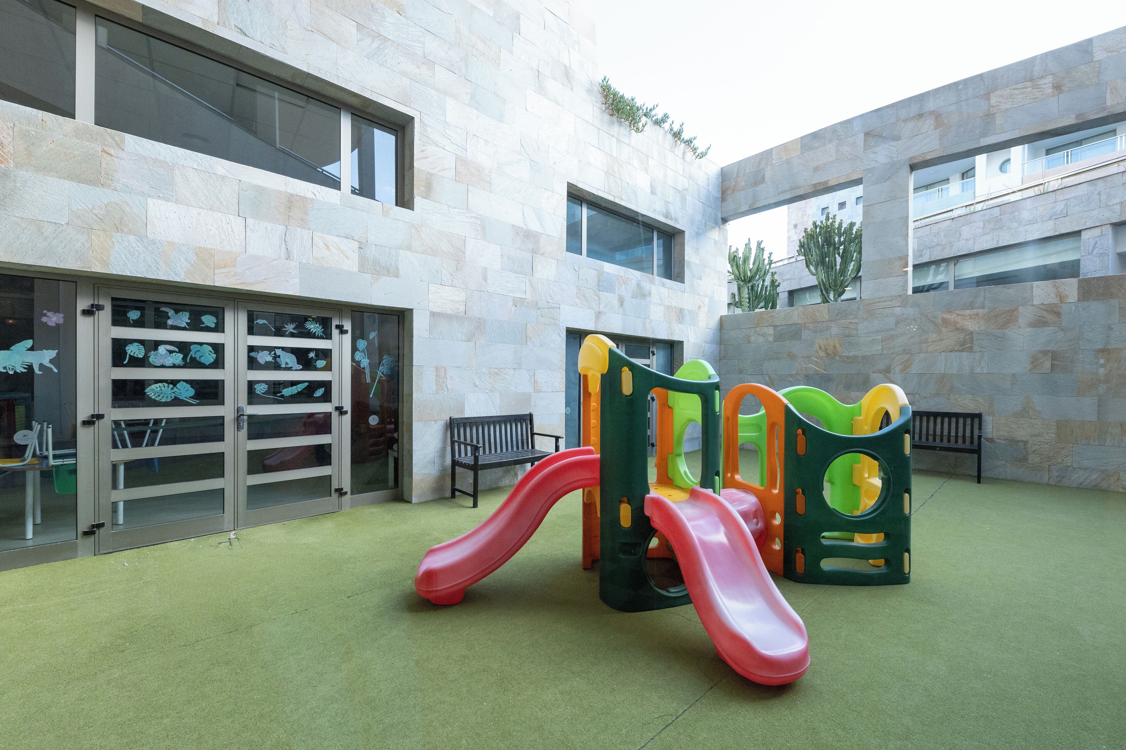 Een speelplaats voor kinderen met glijbanen en klimconstructies, naast een gebouw met grote ramen en een stenen gevel.