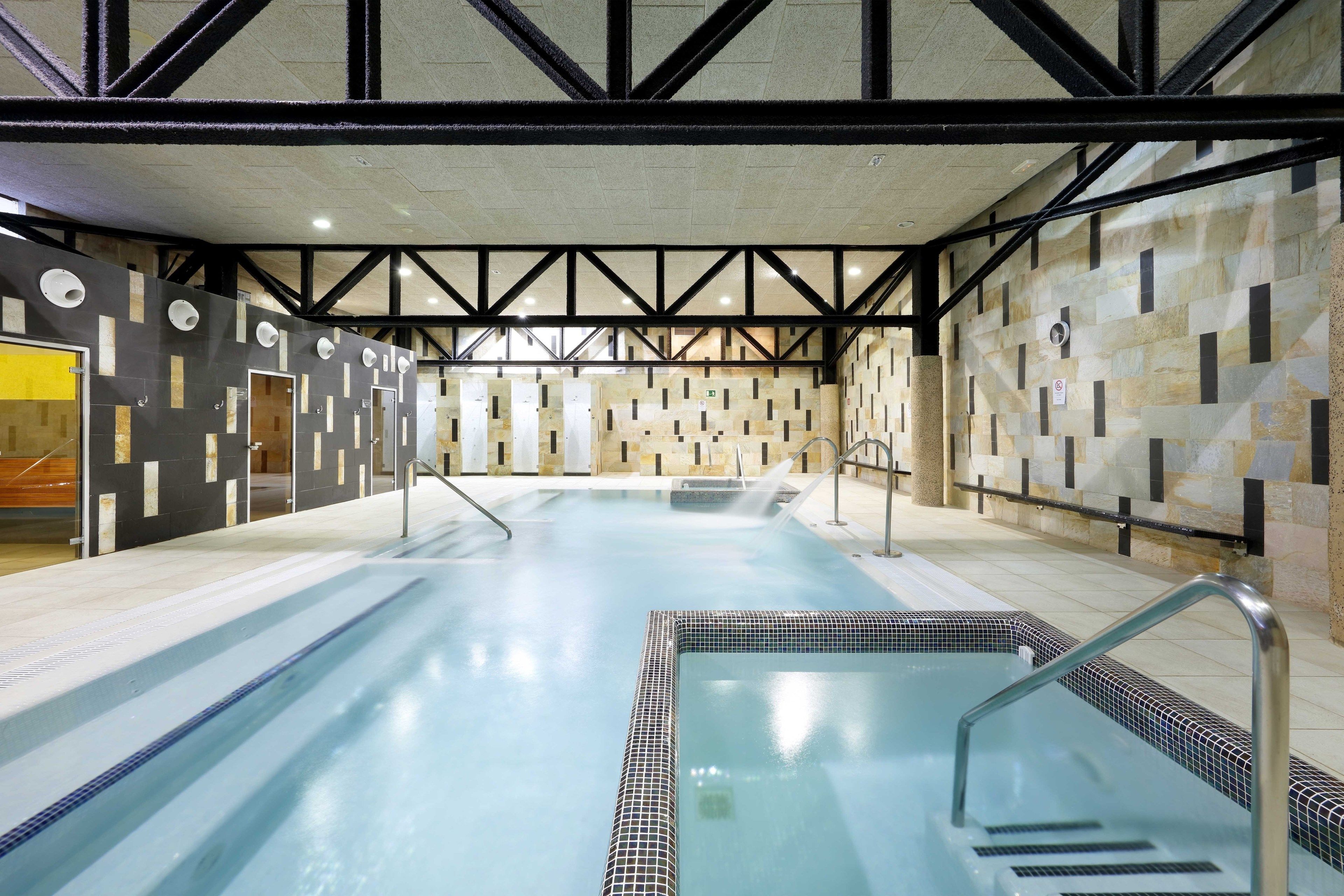 Una piscina amb vora de rajola i una zona poc profunda separada, envoltada per murs de pedra i bigues de metall negre.