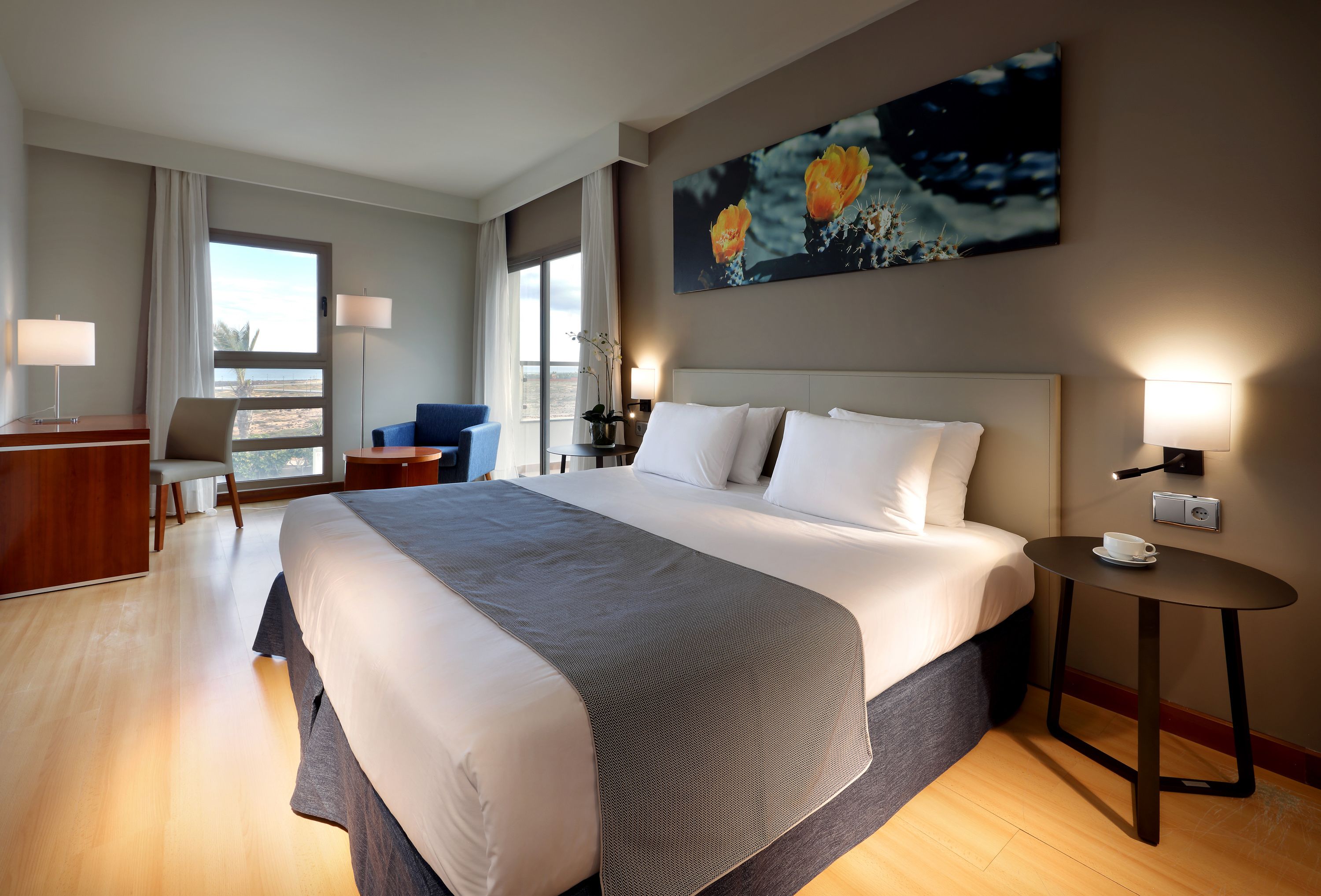A hotel room with a large bed, a desk, a chair, a small table, and a painting on the wall.