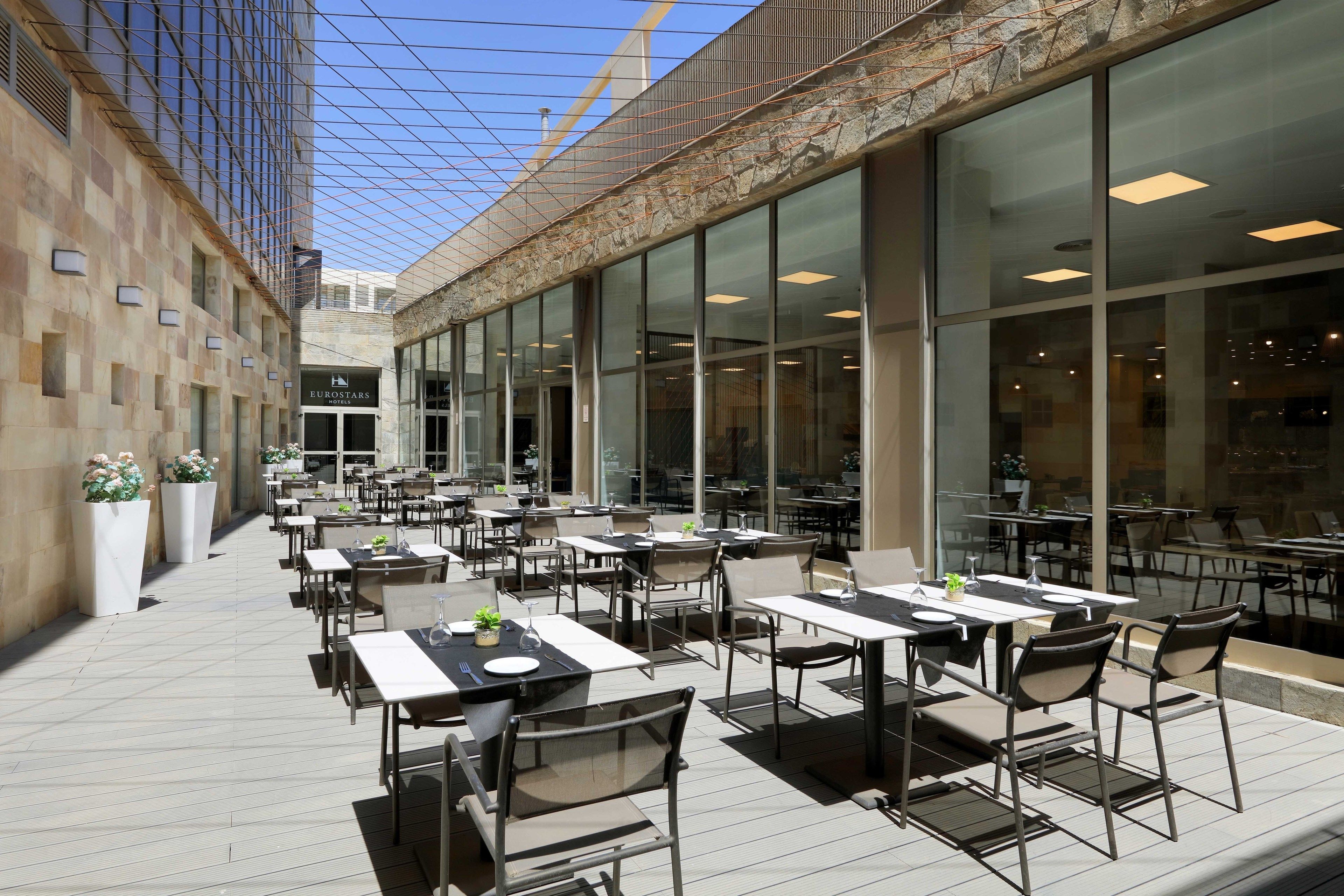 An outdoor dining area with tables and chairs, large windows, and a stone facade.