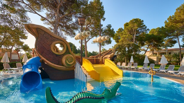 Un área de piscina con tobogán, estructura de juegos acuáticos y sillas de descanso.