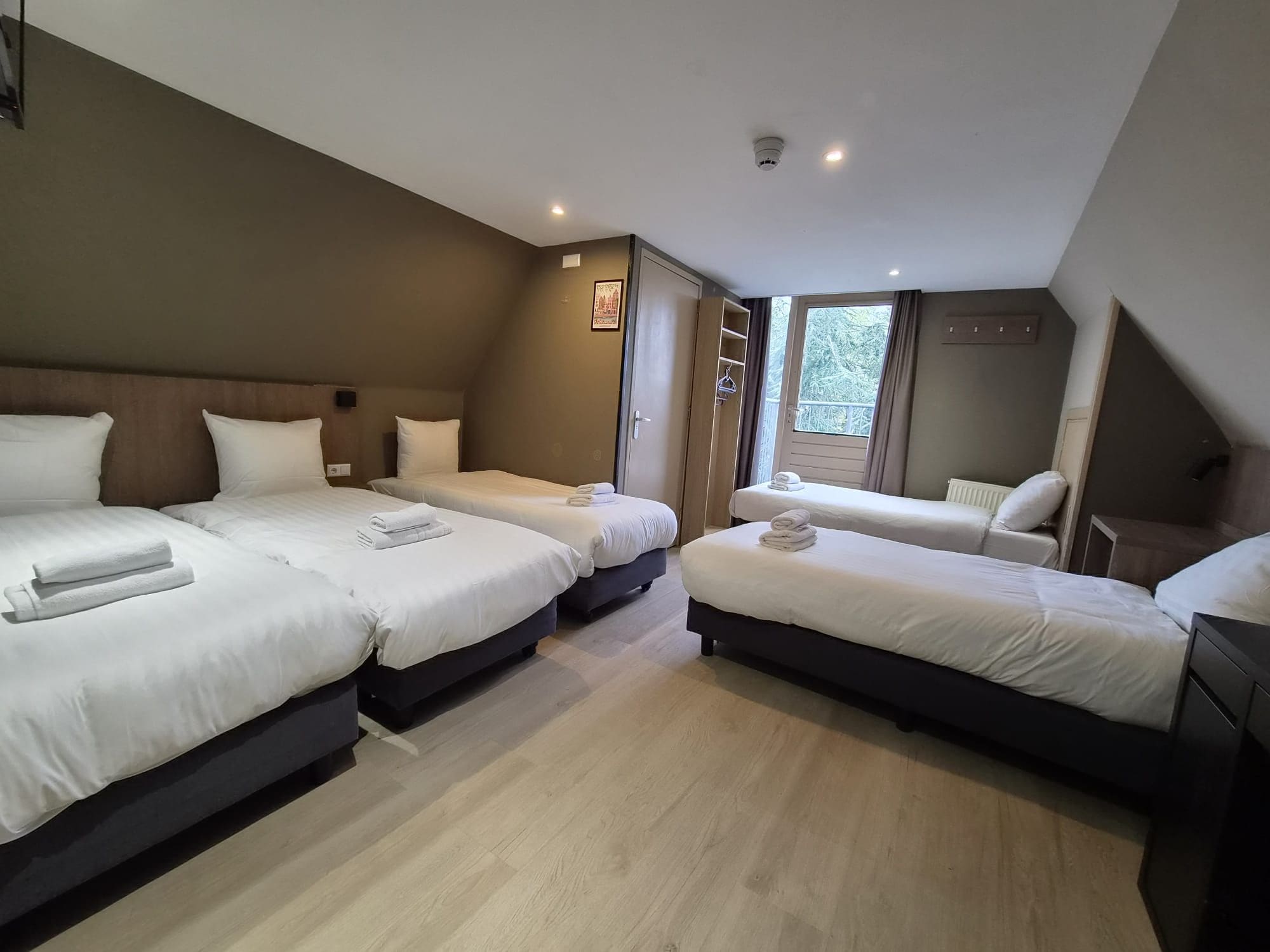 A hotel room with three beds, a wardrobe, a radiator, and a window with a view of trees.