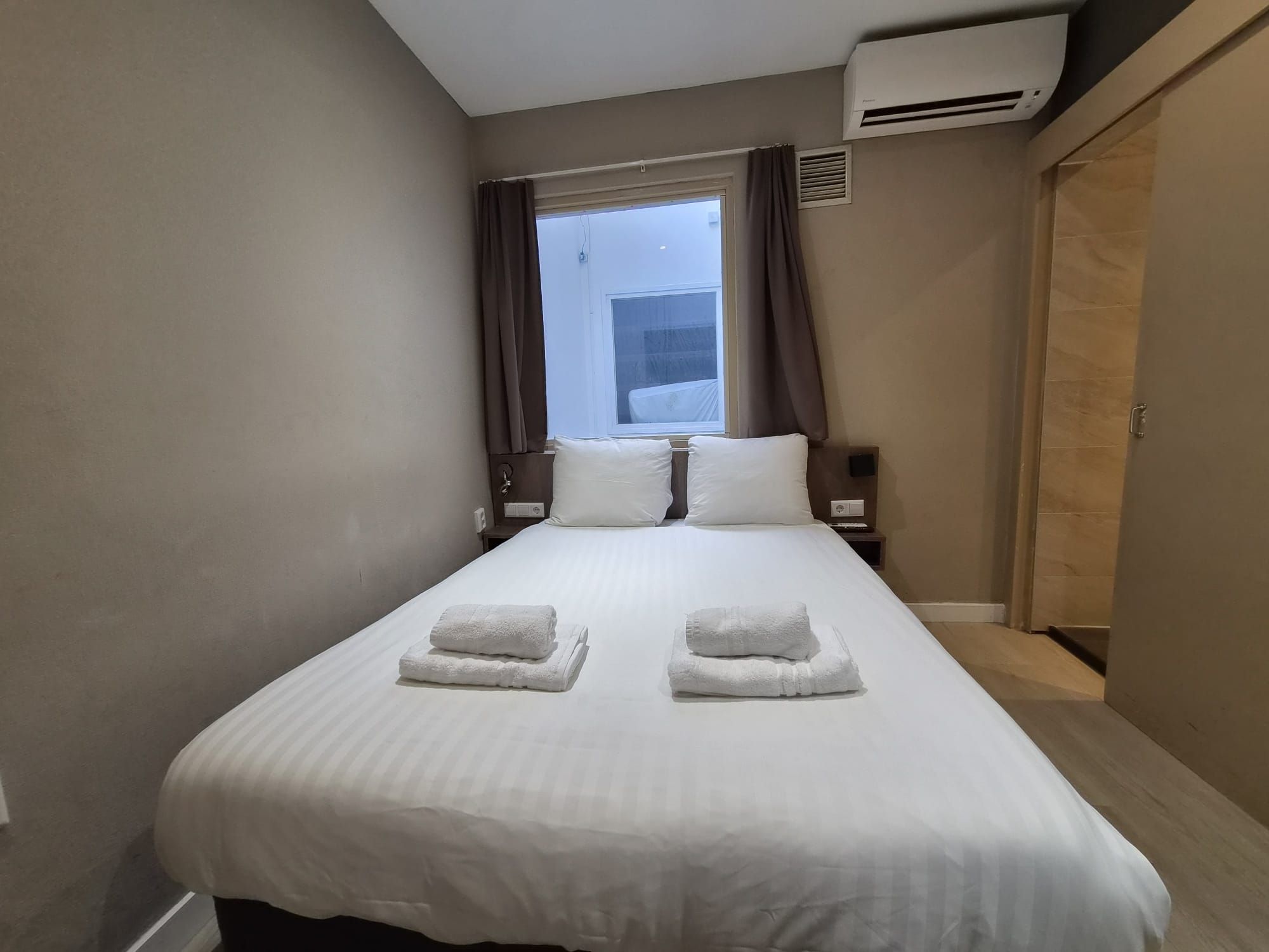 A hotel room with a bed, two pillows, a nightstand, a bedside table, a window with curtains, an air conditioning unit, and a door.