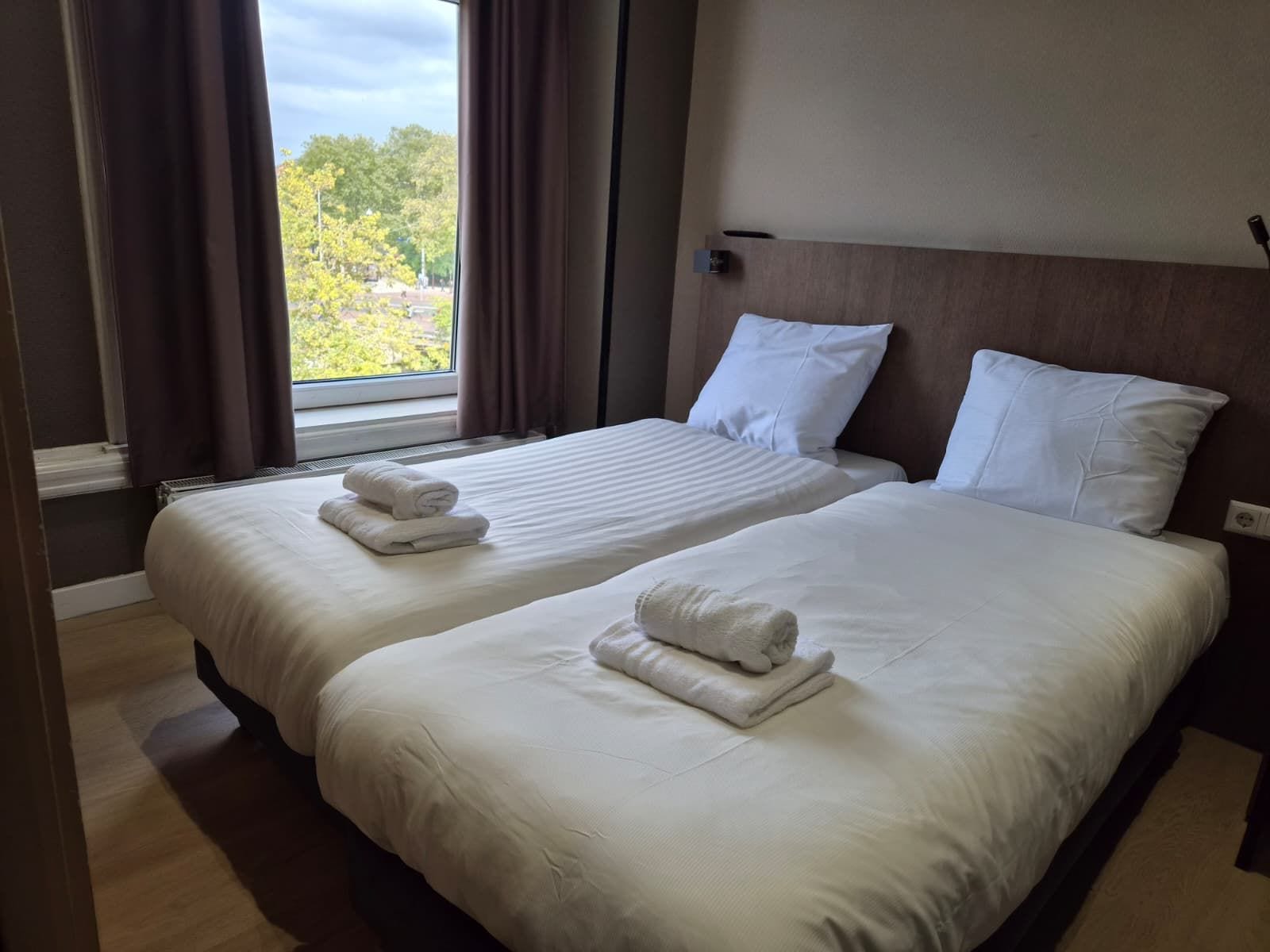 A hotel room with two beds, each with white linens and towels, a window with curtains, and a view of trees outside.