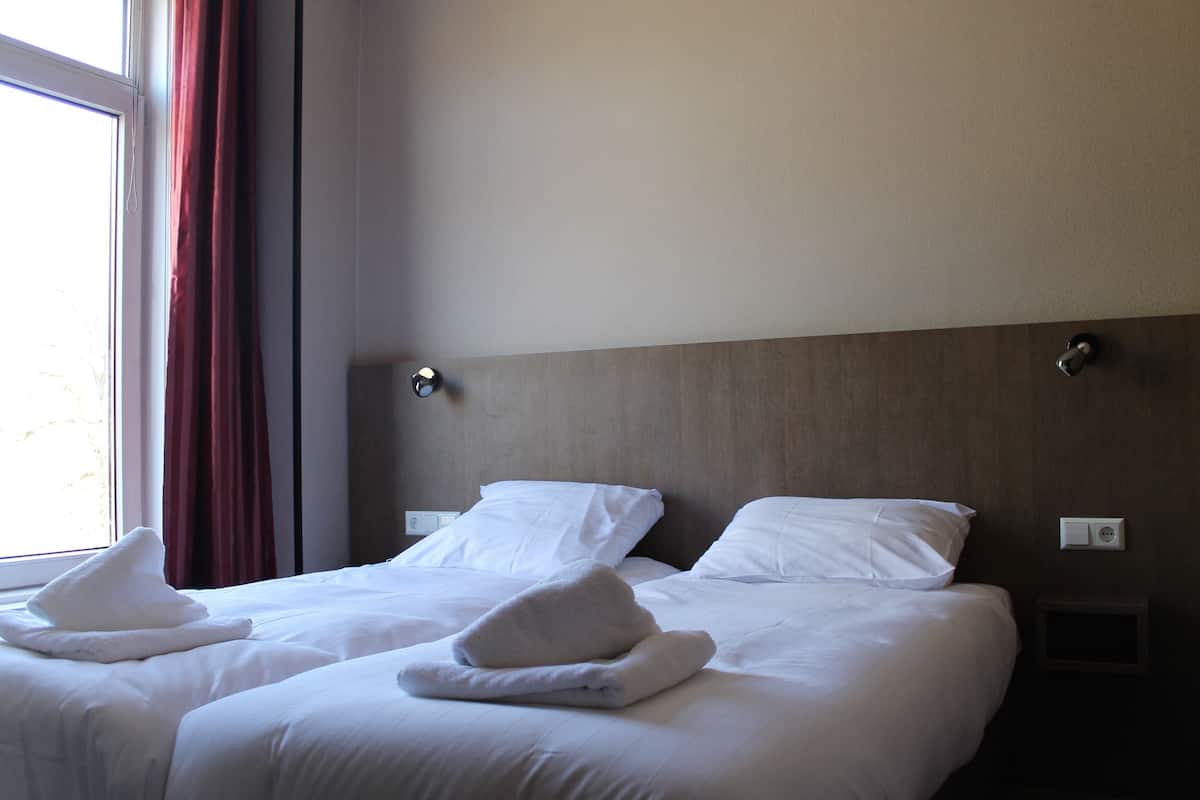 A hotel room with two beds, each with white linens and towels, a wooden headboard, and a window with red curtains.