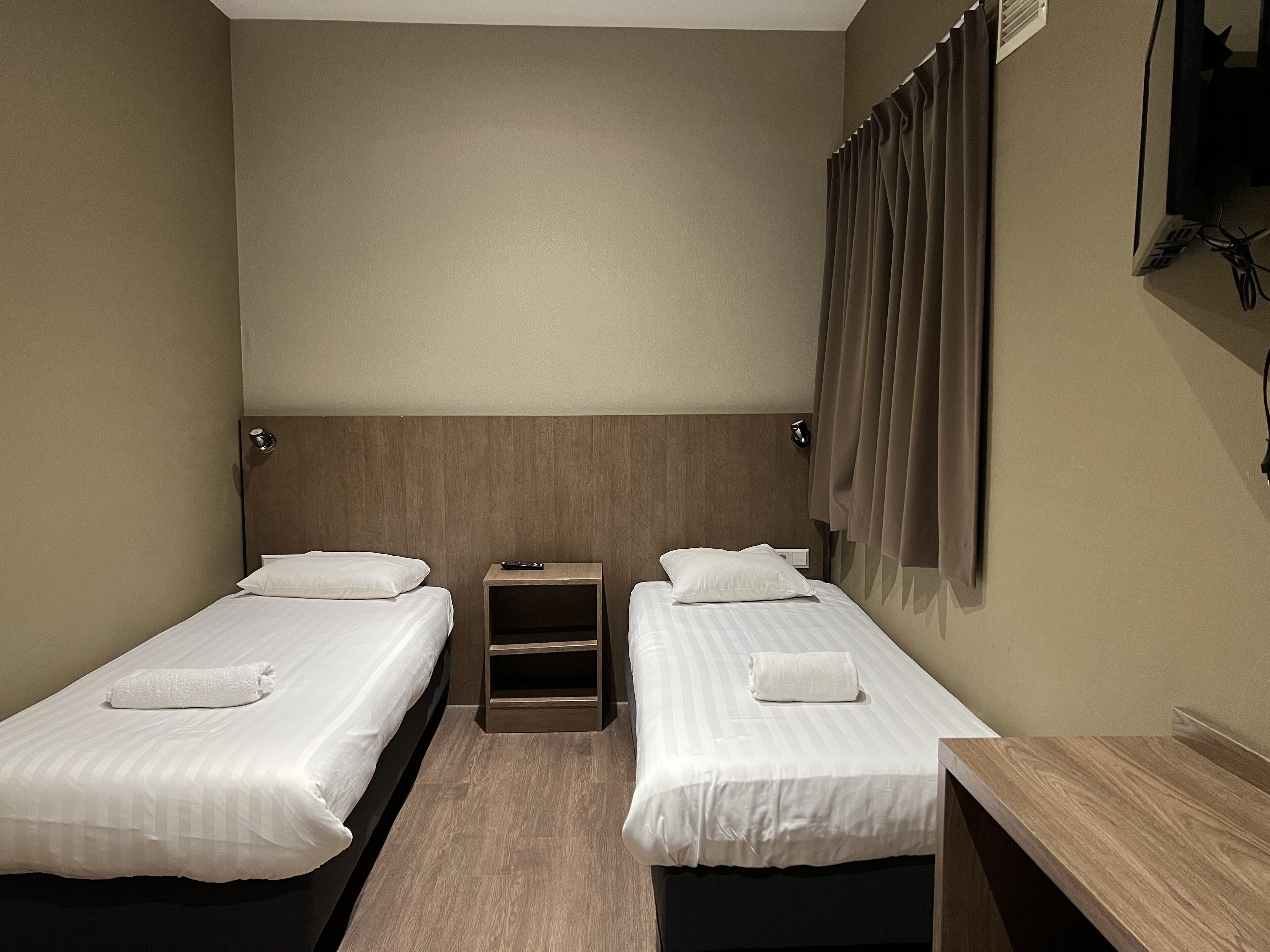 Quarto de hotel com duas camas de solteiro, cabeceira de madeira, mesa de cabeceira, televisão fixada na parede e janela com cortinas.