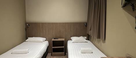 Quarto de hotel com duas camas de solteiro, cabeceira de madeira, mesa de cabeceira, televisão fixada na parede e janela com cortinas.