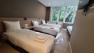 A hotel room with three beds, a flat-screen TV mounted on the wall, and large windows with a view of greenery outside.
