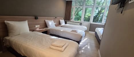 Habitación de hotel con tres camas, un televisor de pantalla plana montado en la pared y ventanas amplias con vistas a zonas verdes.