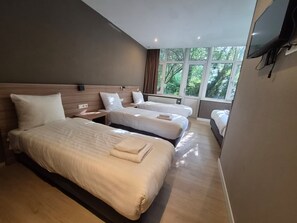 A hotel room with three beds, a flat-screen TV mounted on the wall, and large windows with a view of greenery outside.