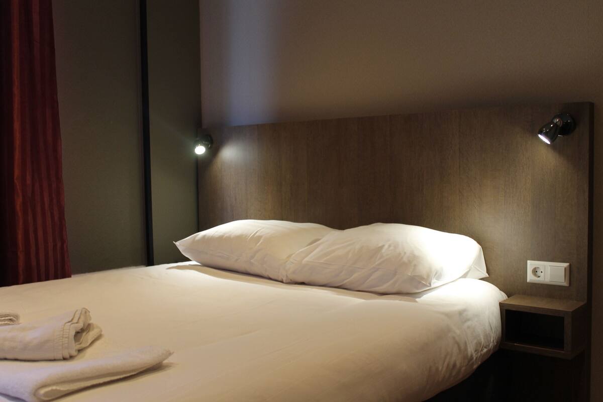 A neatly made bed with white linens and pillows, a bedside lamp, and a power outlet on the nightstand.