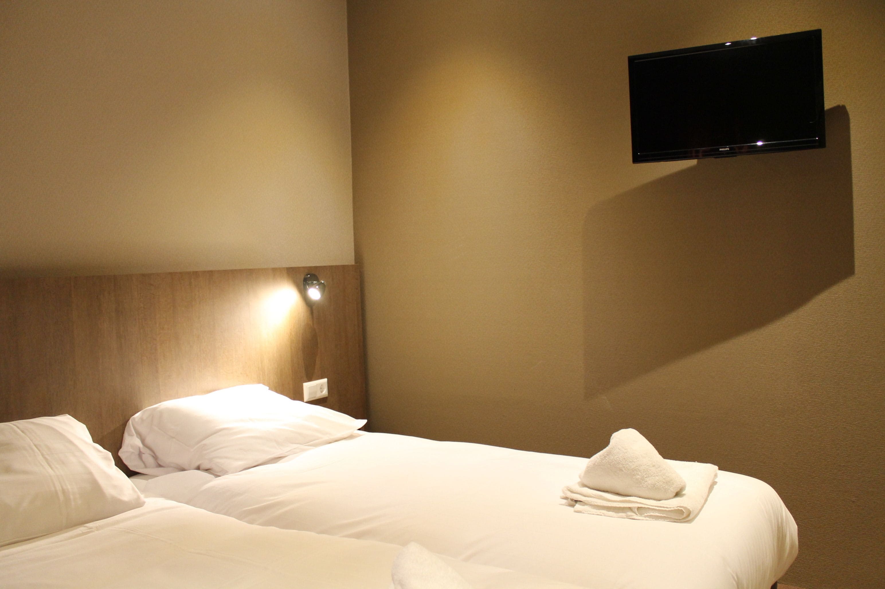 Una habitación de hotel con cama, televisor montado y mesita de noche con lámpara.