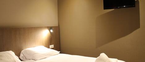 Una habitación de hotel con cama, televisor montado y mesita de noche con lámpara.