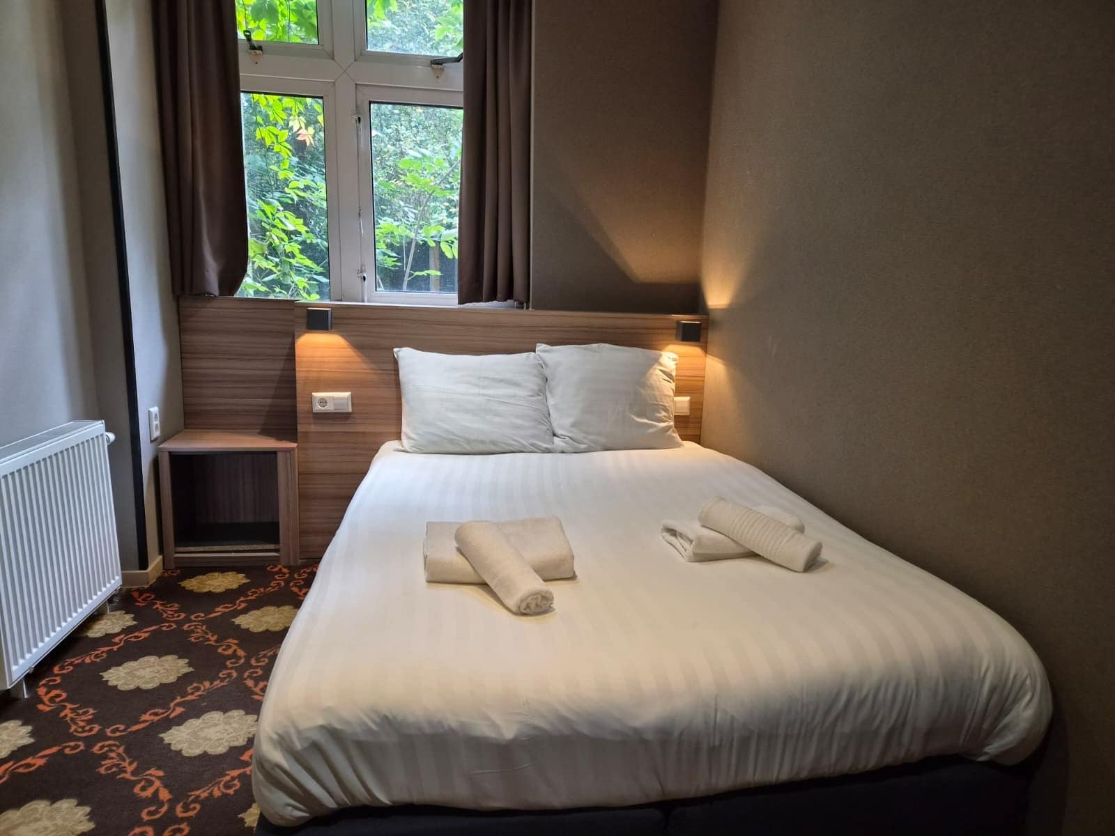 Una habitación de hotel con cama, ventana con cortinas, mesita de noche y radiador.