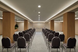 Un salón de conferencias con filas de sillas frente a una larga mesa, paneles de madera en las paredes e iluminación empotrada en el techo.